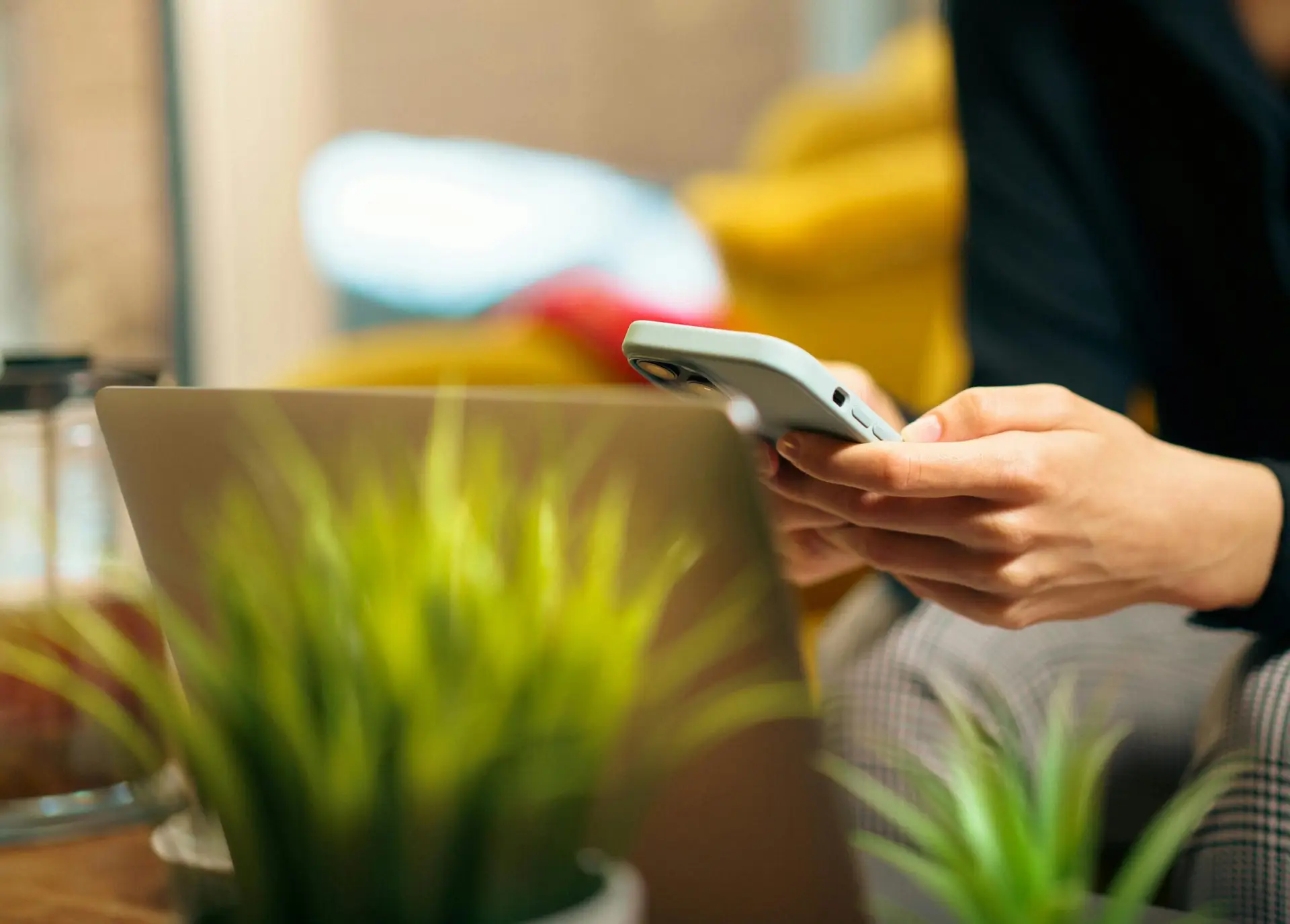 A woman multitasking on her smartphone and laptop surrounded by green plants in a cozy living room.